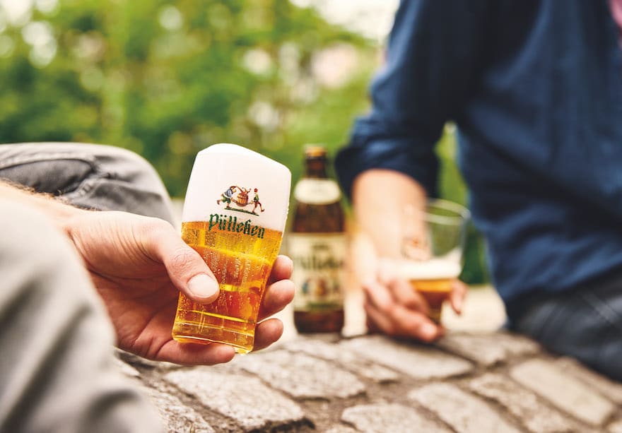 Mann hält Glas mit Markenlogo "Helles Pülleken" in der Hand