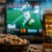 TV-Bildschirm mit Fußballspiel in einem Wohnzimmer, im Vordergrund Bierglas und Erdnüsse