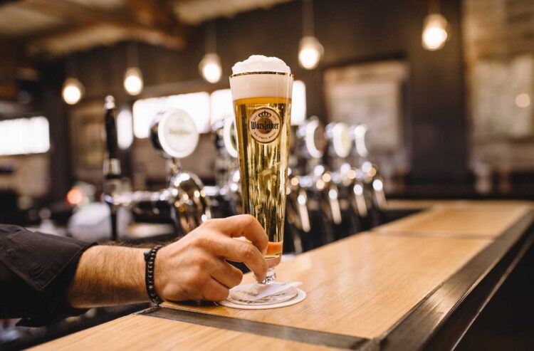 Ein Mann stellt ein Glas Warsteiner auf eine Kneipentheke.