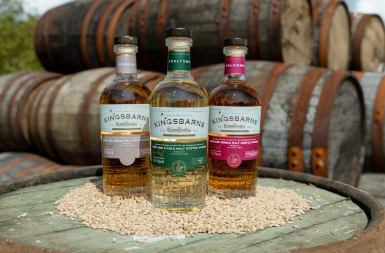 drei verschiedenen Flaschen Kingsbarns, im Hintergrund Whiskyfässer